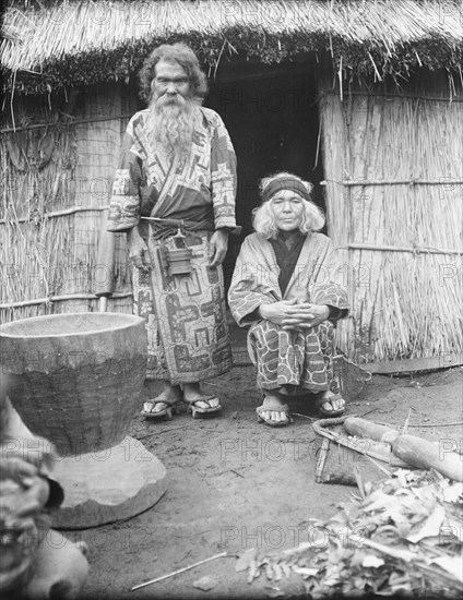 Ainu man and seated woman at the entrance of a hut, 1908. Creator: Arnold Genthe.