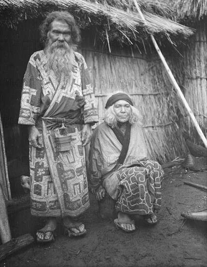 Ainu man and seated woman at the entrance of a hut, 1908. Creator: Arnold Genthe.