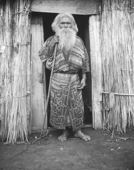 Ainu man holding a staff standing at the doorway of a hut, 1908. Creator: Arnold Genthe.