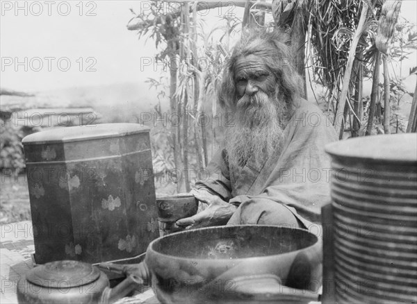 Ainu man seated outdoors on a mat covered with clay containers, 1908. Creator: Arnold Genthe.