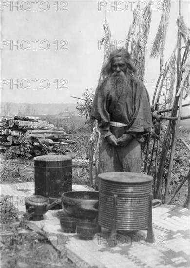 Ainu man standing outdoors on a mat covered with clay containers, 1908. Creator: Arnold Genthe.