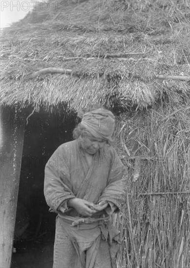 Ainu woman standing outside the entrance of a hut, 1908. Creator: Arnold Genthe.