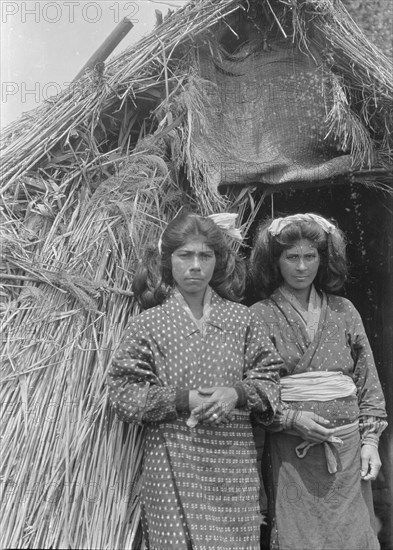 Two Ainu women standing at the entrance of a hut, 1908. Creator: Arnold Genthe.