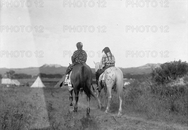 Two daughters of a chief on horseback, riding away from camera toward tents in background, c1907. Creator: Edward Sheriff Curtis.