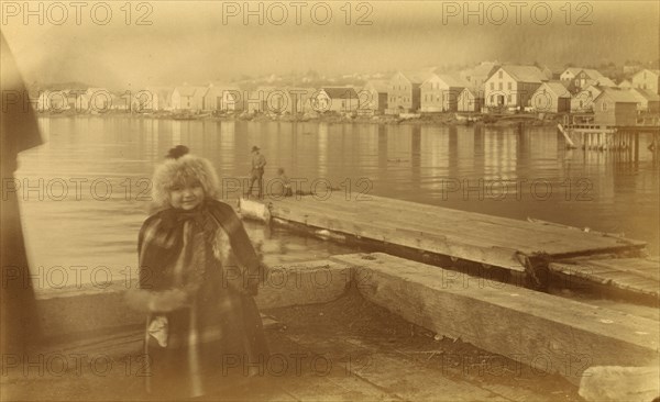 Unidentified young girl standing on wharf, with buildings along waterfront...,1894 or 1895. Creator: Alfred Lee Broadbent.