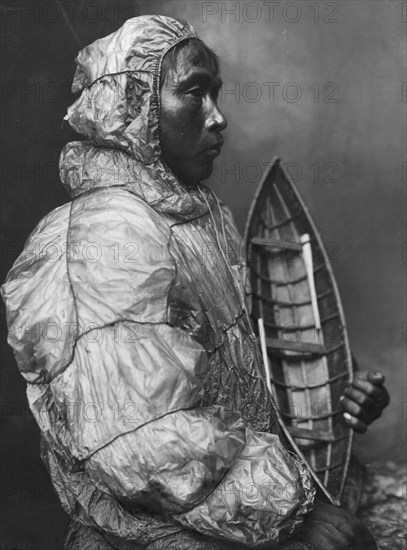 Kayaker wearing water-proof jacket with toy boat he made for his son, between c1900 and c1930. Creator: Lomen Brothers.