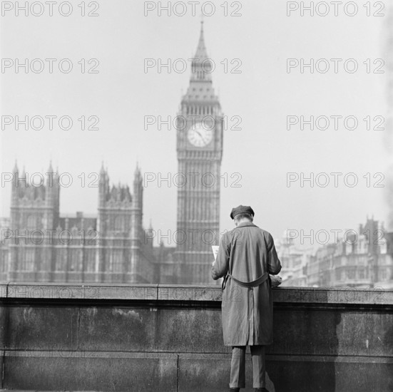 Houses of Parliament seen from the Albert Embankment with a man reading a newspaper by the parapet in the foreground, London, 1964.
