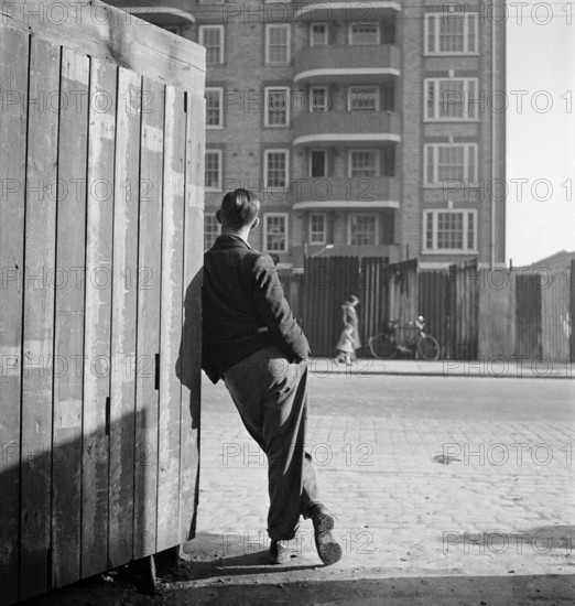 A man leaning on a street corner hoarding looks across the road to where a woman and child pass corrugated iron sheets enclosing an apartment block, possibly Rugmere on Ferdinand Street, Camden, 1945-1950.