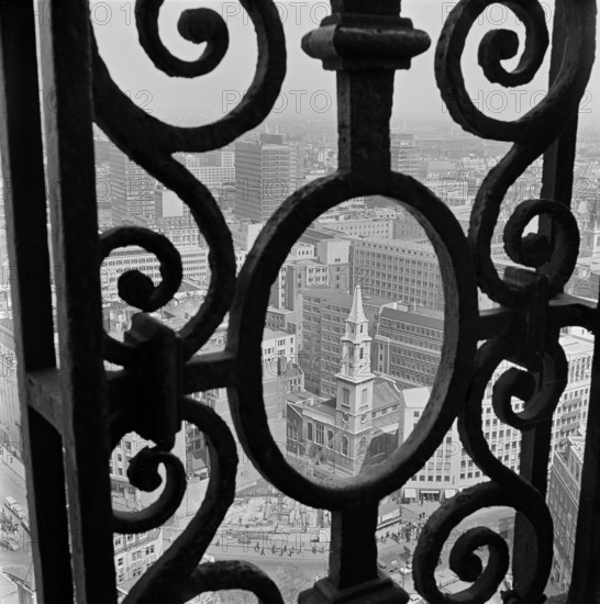 View through the decorative iron railings of the Golden Gallery at St Paul's Cathedral, looking north-east over the rooftops of the surrounding buildings, with the tower of St Vedast's Church in the centre of the frame, 1960-1970.
