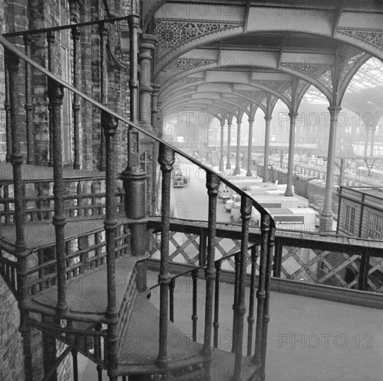 Interior of Liverpool Street Station showing a cast iron spiral staircase, 1960-1972. Visible in distance are platforms and freight area with trucks.