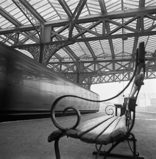 Looking along a bench with curved wrought iron arms on a platform at Waterloo Station with a train passing at speed in the background, 1960-1972.