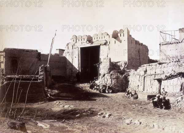 Lahore Gate, Kabul, Afghanistan, c1879. Small groups of people sit along the wall. A dirt track leads up to the gate which is in a delapidated state and falling into ruin.
