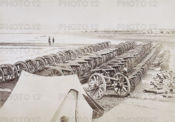 Captured guns, Kabul, Afghanistan, c1879. Cannons lined up in neat rows. A British soldier leans against a wheel for scale. On the right is a pile of dismantled guns. A camp can just be seen in the distance on the right.