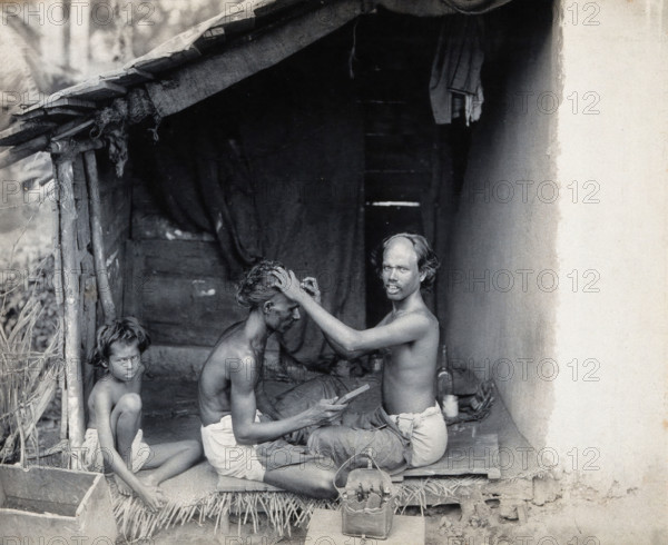 Two men sitting cross-legged; one is a barber shaving the front of the other's head, c1900. Creator: Unknown.