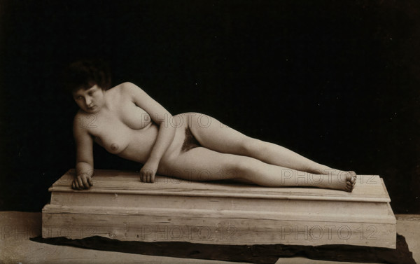 A young woman posing naked in a photographic studio, lying on a wooden plinth, approx 1900. Creator: Unknown.