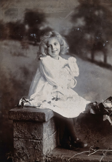 A girl, holding a doll, sitting on a wall, c1890s. Creator: Lock & Whitfield.