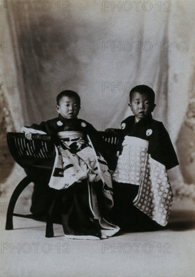 Two boys in a photographic studio, one seated on a chair, in front of a curtained backdrop, c1900. Creator: Unknown.