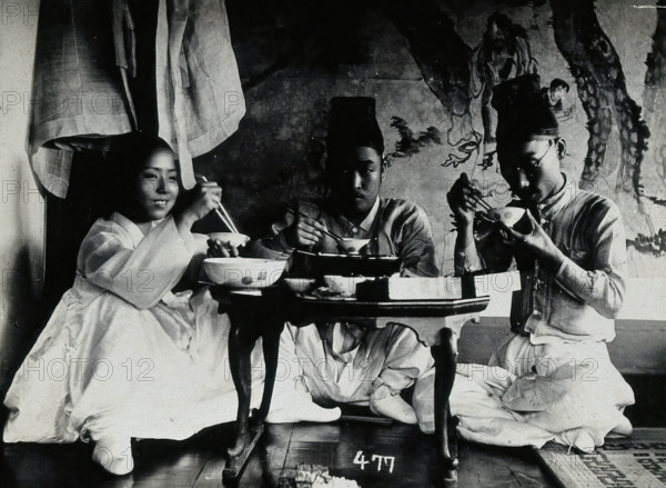 A group of people eating a meal, seated behind a small table, in front of a painted screen, c1900. Creator: Unknown.