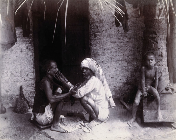 Two Indian men crouching; one is cutting the other's hair, while a girl sits.., c1890s. Creator: Unknown.