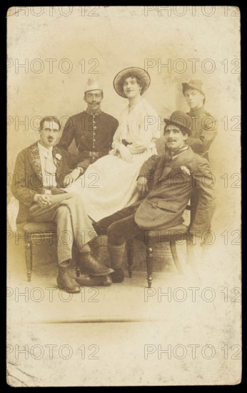 Amateur actors sitting down, surrounding a man in drag seated in the centre, [1910-1919?]. Creator: Unknown.