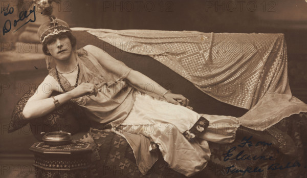 A man in drag poses wearing delicate attire; curled up on a piece of furniture, [1910-1919?]. Creator: Unknown.