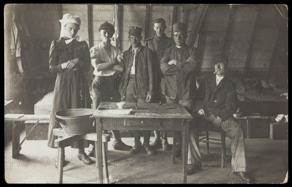 French prisoners of war, one in drag, gather round a small wooden table, [between 1910 and 1919?]. Creator: Unknown.