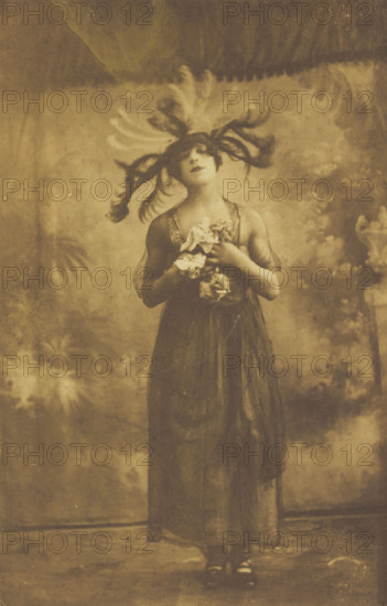 A performer in drag poses on stage wearing a head garment with sprawling feathers, [1910-1919?]. Creator: Unknown.
