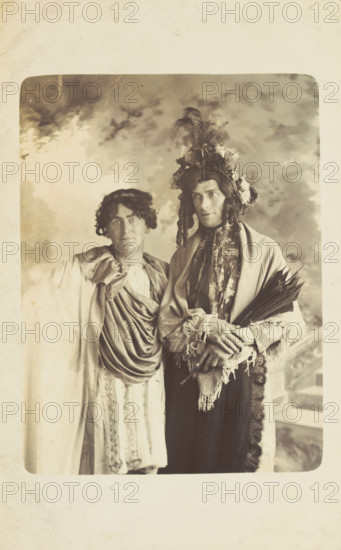 Two men in drag, one wearing a detailed feathered head garment, pose for a portrait, [1900-1909?]. Creator: Frank Wood.