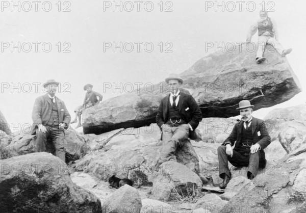 Unknown men and boy, unknown location posing on large rocks, c1900s. Creator: Robert Augustus Henry L'Estrange.