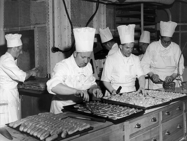 En juillet 1949, à bord du paquebot de luxe L'ILE DE FRANCE, des pâtissiers s'affairent à la tâche devant des plateaux garnis de succulentes patisseries.