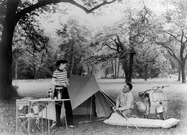 En 1955, un couple campe au milieu d'un champ. Ils disposent de tous les objets qui caractérisent le camping MODERNE : la table et les chaises pliantes, le réchaud à gaz, la tente deux places et le matelas pneumatique. Pour se déplacer, ils utilisent un nouveau véhicule deux roues, le scooter VESPA .