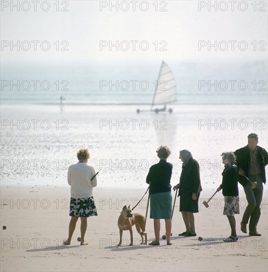 Dans les années 1970, sur une plage normande, des vacanciers jouent au croquet en regardant passer les chars à voile.