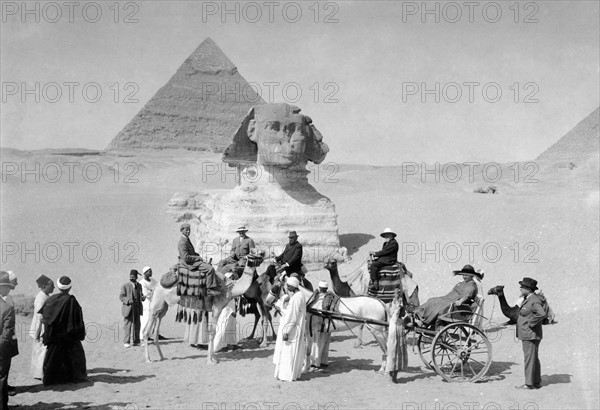Début du 20e siècle, devant le Sphinx de Gizeh dans la Vallée des Rois (Basse-Egypte), un groupe de touristes européens montant des chameaux ou assis dans une carriole. Sculpté à l'effigie du pharaon Khephren, l'animal à tête humaine garde la pyramide de ce dernier située à l'arrière. La pyramide de Khephren est la seule des trois pyramides monumentales de Gizeh à avoir conservé son revêtement intact dans sa partie supérieure. L'ensablement de la Vallée des Rois est alors tel que les pattes du Sphinx sont complètement recouvertes.  Cliché de Jean Clair-Guyot, inédit du fonds L'Illustration.