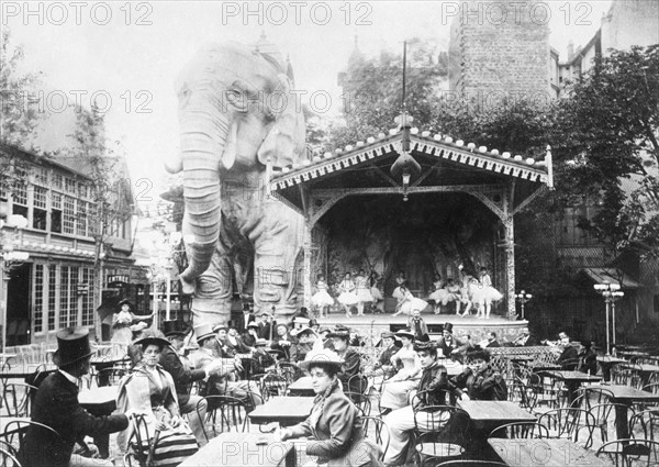 Vers 1880, à Paris, consommateurs assis dans le jardin intérieur du Moulin-Rouge, transformé en café-concert pendant la saison estivale.  A gauche de la scène où se produisent les danseuses, un gigantesque éléphant de stuc, vestige de l'Exposition universelle de 1889.
