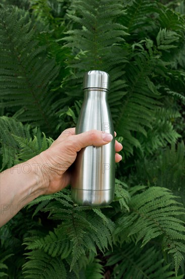 Reusable metal water bottle in female hand to replenish the body's water reserves against the background of fern leaves in tropics.