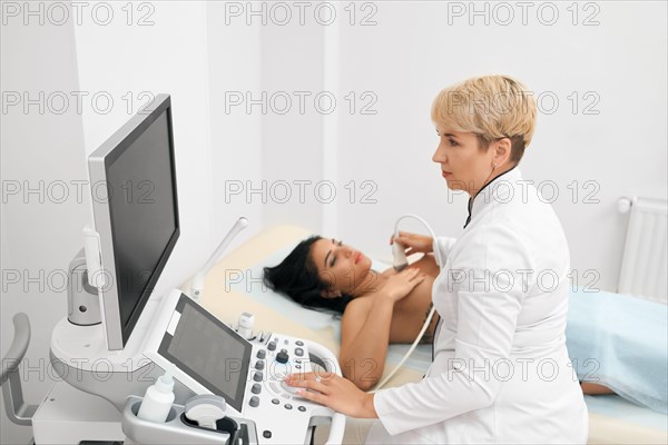 Concentrated doctor in white medical uniform looking at monitor from uzi apparatus while performing breast examination to young female patient. Concept of breast cancer prevention