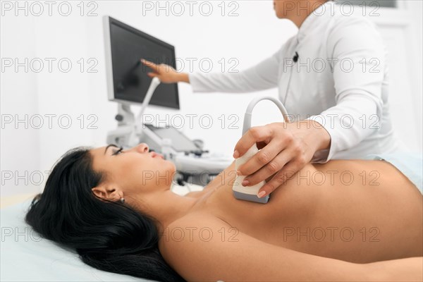 Attractive woman with dark hair lying on couch with naked breast while competent female doctor doing ultrasound diagnostic at modern clinic. Concept of breast cancer prevention