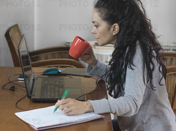 Brunette woman telecommuting at home for COVID-19, looking at the computer and drinking coffee