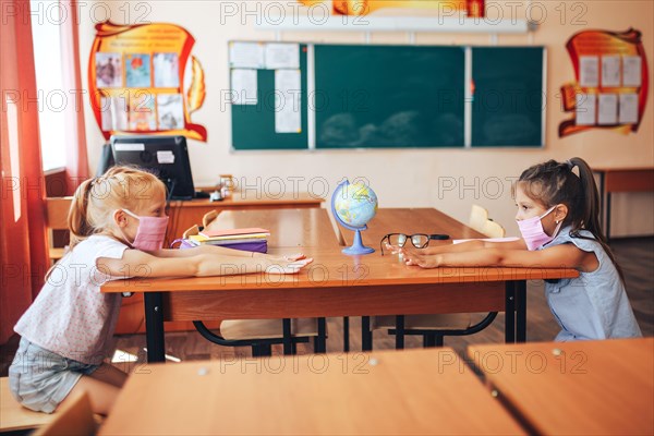 Two schoolgirls in medical masks are sitting at a school desk, opposite each other, group session, back to school, teaching children, social distance