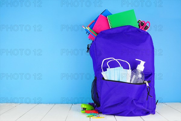 Backpack full of school supplies and COVID 19 prevention supplies. Blue background. Back to school during pandemic concept.