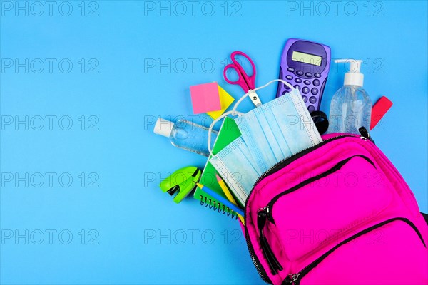 Backpack full of school supplies and COVID 19 prevention items. Top view, spilling onto a blue background. Back to school during pandemic concept.