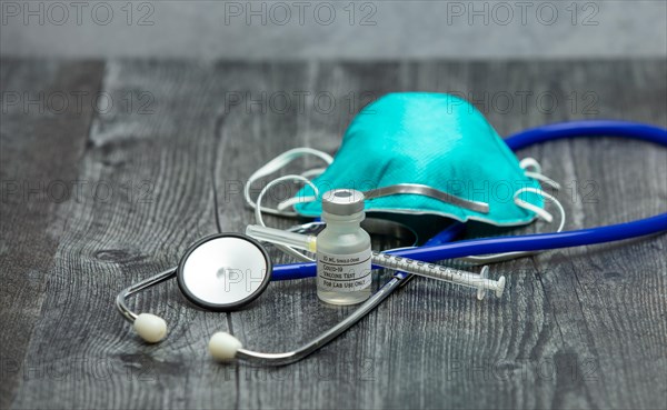 A blue medical stethoscope, face mask respirator, syringe and vial of covid-19 test vaccine on a wooden surface.