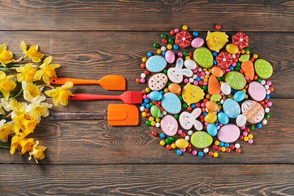 Colorful ginger glazed cookies, daffodils and chocolate balls with sugar shell. Homemade lovely pastry of easter animals, eggs and carrots in shape of circle, three silicone kitchen tools nearby.