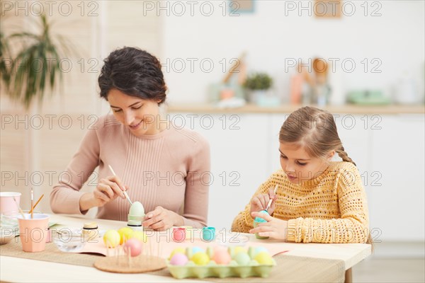 Candid portrait of mother and daughter painting Easter eggs pastel colors sitting at table in cozy kitchen interior, copy space