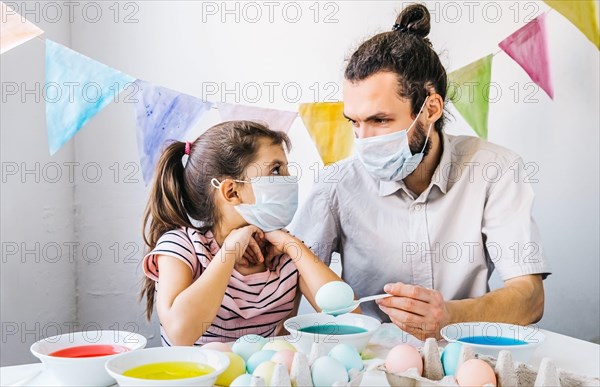 New normal Easter celebration lifestyle concept. Cute little girl and her dad wearing protective masks, preparing easter, dyed eggs at home during