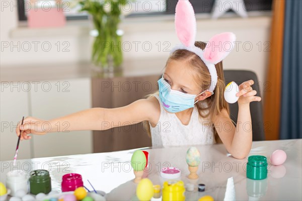 Pretty little girl wearing bunny ears and disposable medical face mask and painting easter eggs at home during coronavirus covid-19 outbreak.