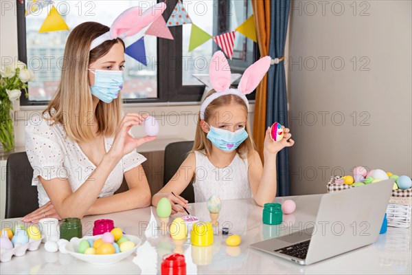 Pretty little girl wearing bunny ears and disposable medical face mask and painting easter eggs at home during coronavirus covid-19 outbreak.