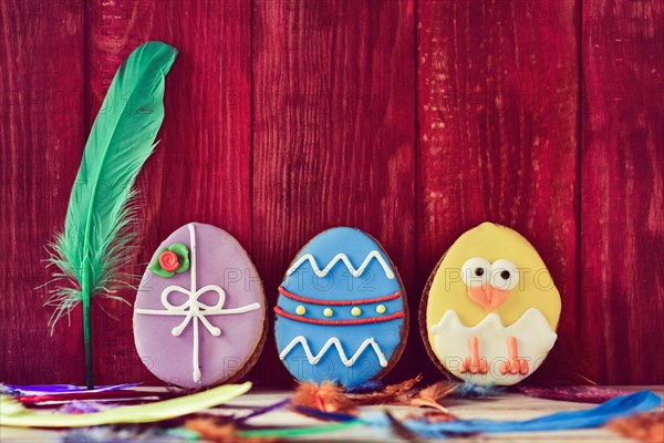 a pile of different some cookies patterned as different decorated easter eggs against a red wooden background, and feathers of different colors sprink