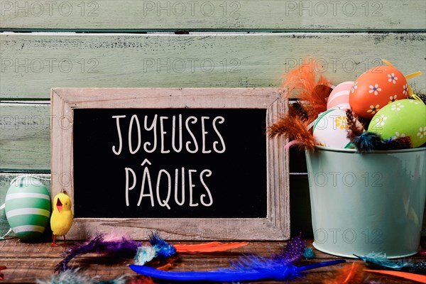 a chalkboard with the text joyeuses paques, happy easter in French, hanging on a rustic wooden wall, and a pile of different decorated easter eggs, a
