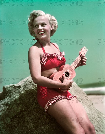1940s 1950s SMILING BLOND WOMAN WEARING RED BIKINI BATHING SUIT LEANING ON ROCK AT BEACH PLAYING UKULELE